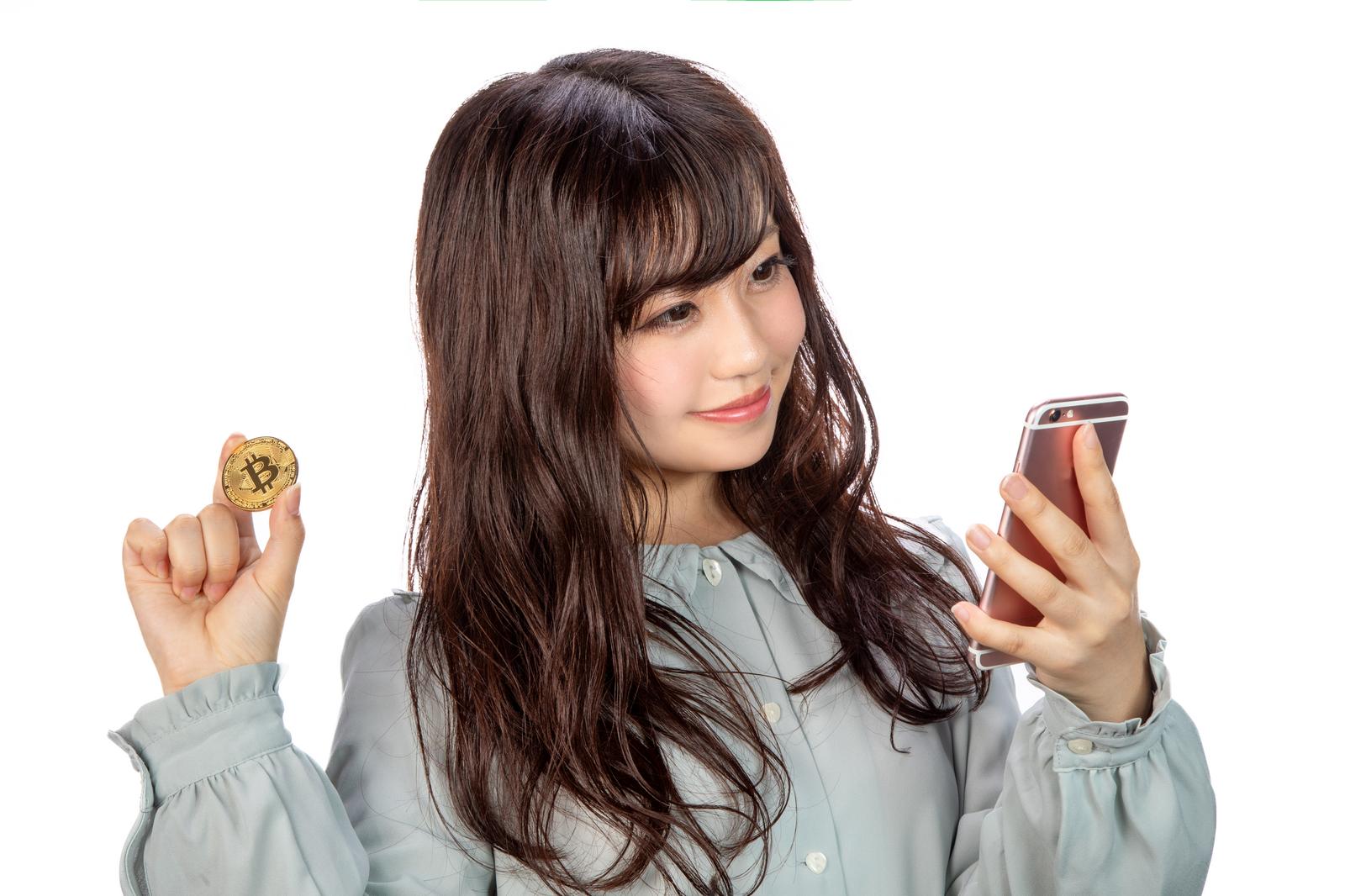 ビットコインを持ってスマートフォンでトレードする女性［モデル：河村友歌］