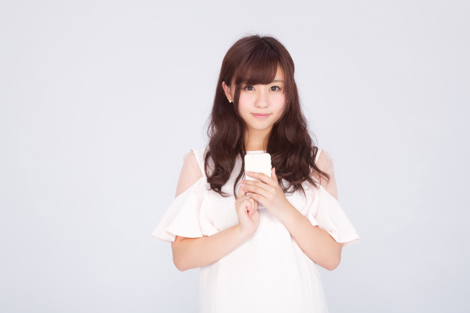 白い服を着たロングヘアの女性が両手でスマートフォンを持っている［モデル：河村友歌］