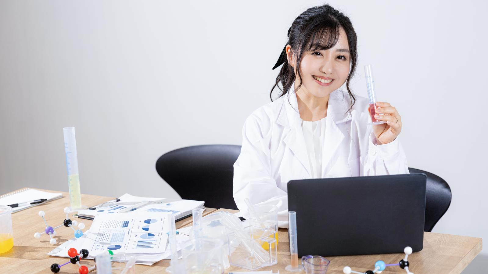 試験管を手に持ち笑顔を見せる白衣姿の女性研究員と、分子模型や資料が並ぶデスク［モデル：河村友歌］