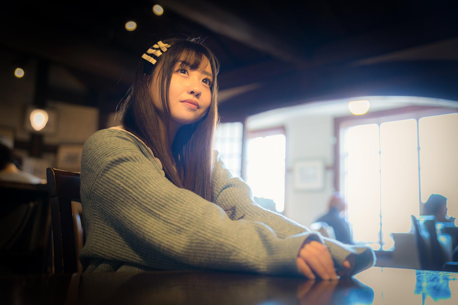 A woman in a sweater sits at a cafe table with her arms resting on it, looking upward while bathed in window light