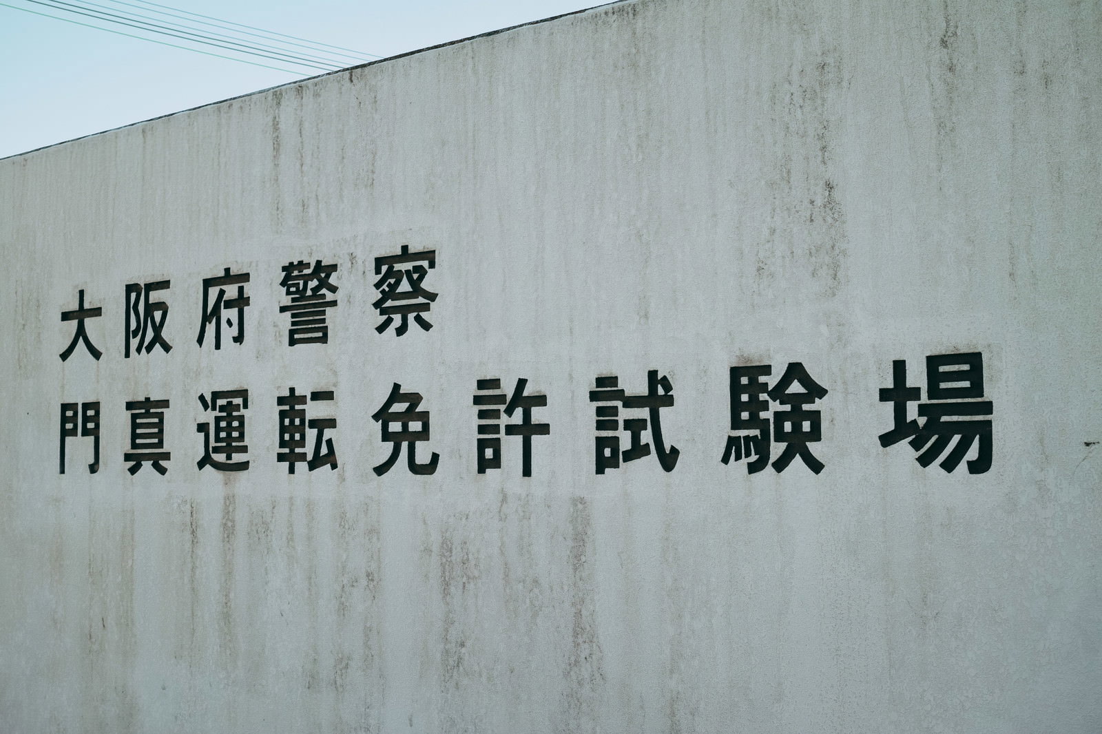 コンクリート壁に黒文字で「大阪府警察門真運転免許試験場」と書かれた看板