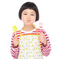 エプロン姿でご飯を待つ女の子がスプーンとフォークを持って食卓に向かうの写真