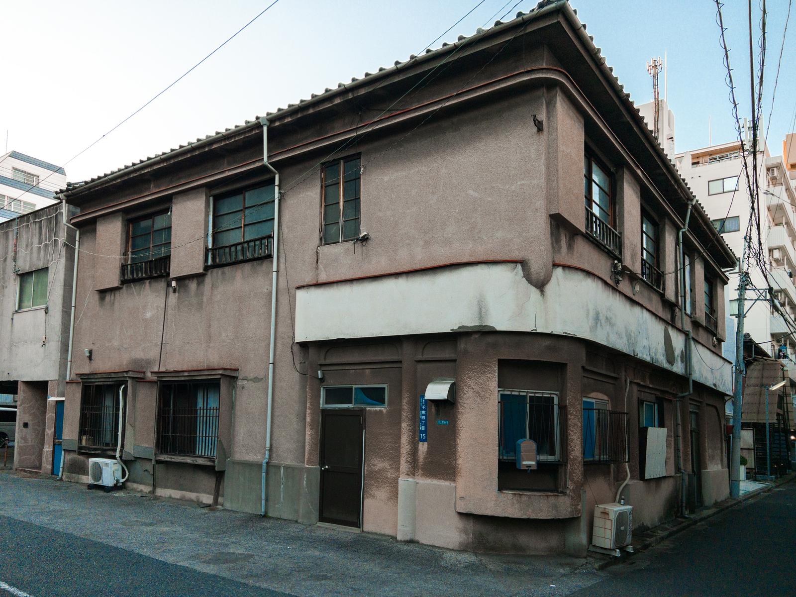 台東区千束の古い建物。グレーベージュの外壁に瓦屋根、複数の窓と木製手すりが見える