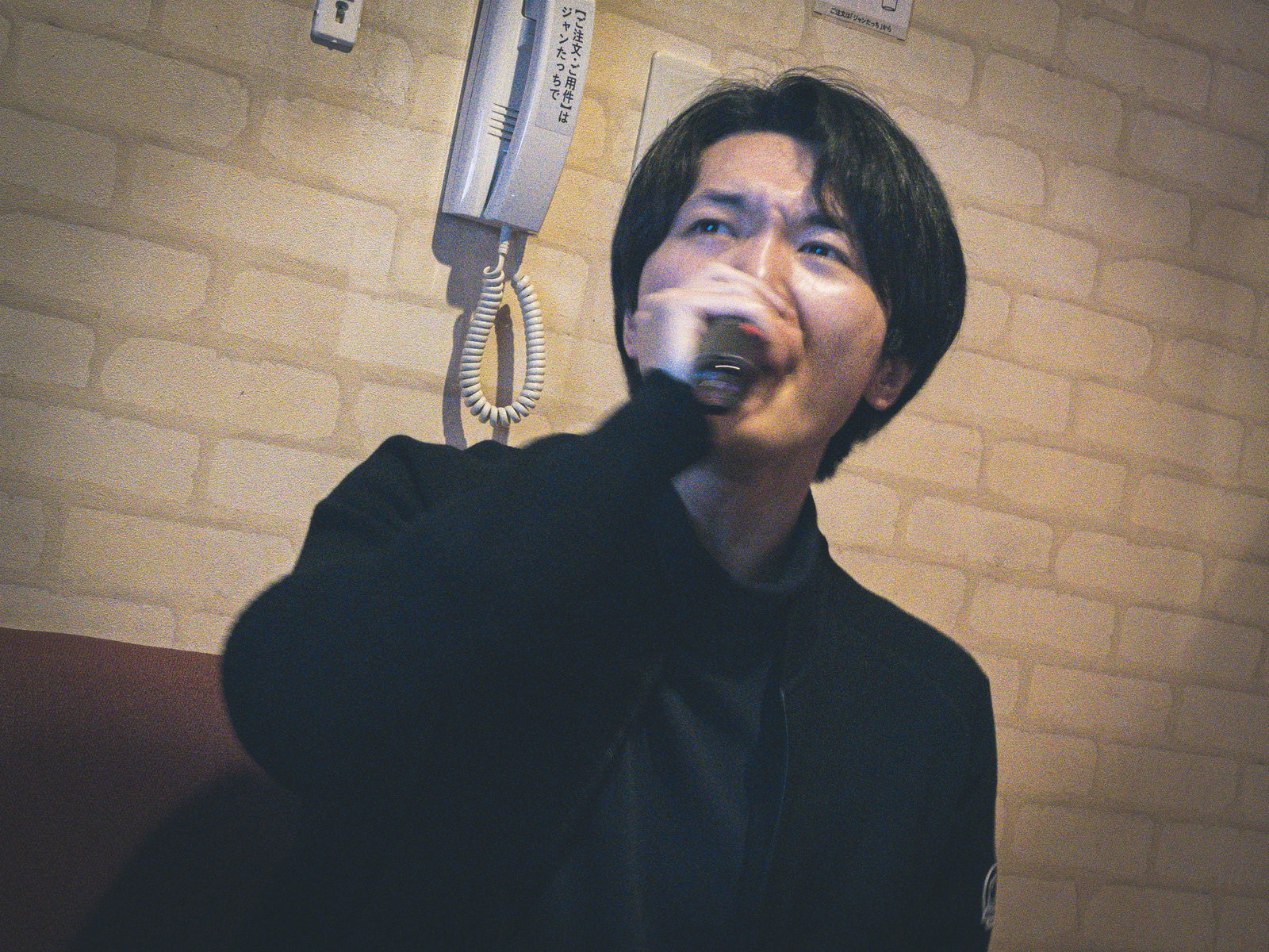 カラオケボックスでマイクを口元に当てて熱唱する黒い服の男性［モデル：花和瞭］