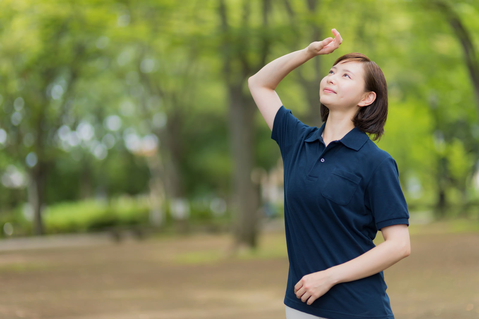 森林の公園で木漏れ日を見上げる紺色制服の介護士女性［モデル：yumiko］