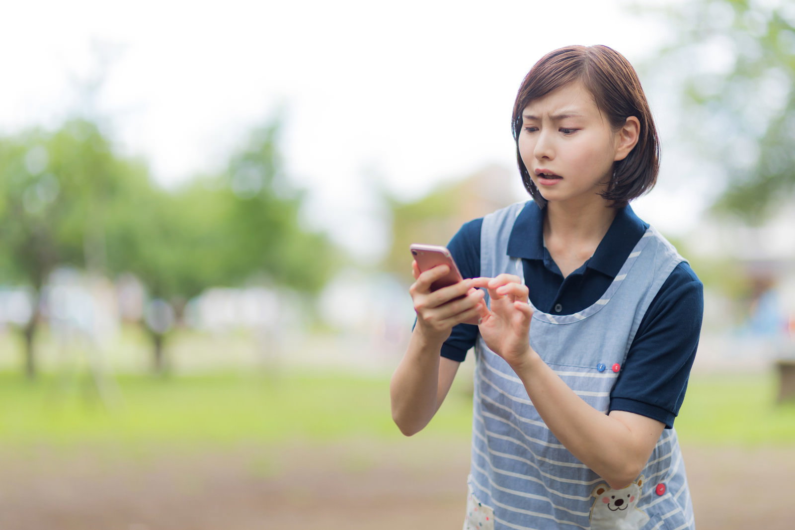 エプロン姿の女性介護士が屋外でスマートフォンを見つめている［モデル：yumiko］