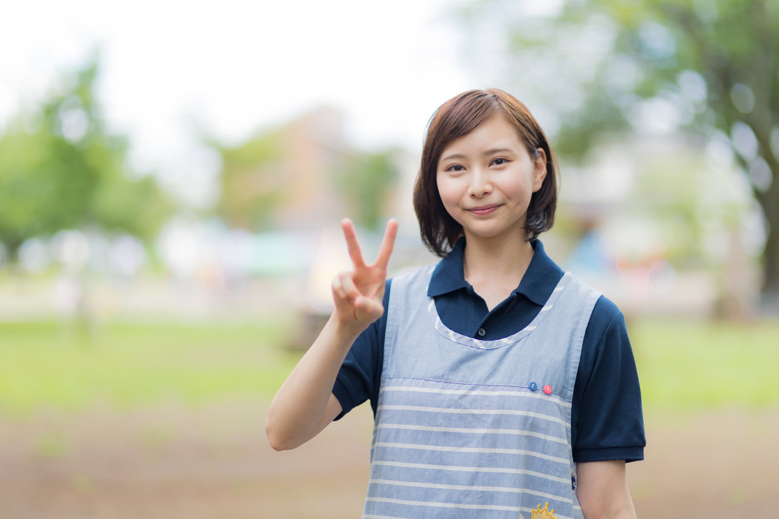 ピースサインをする介護士の女性、笑顔で制服とエプロン着用［モデル：yumiko］