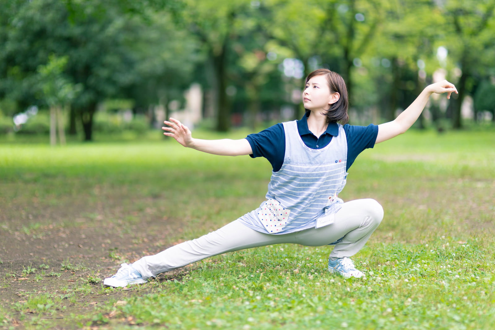 緑豊かな公園で太極拳のようなポーズを取る白いウェア姿の女性介護士［モデル：yumiko］