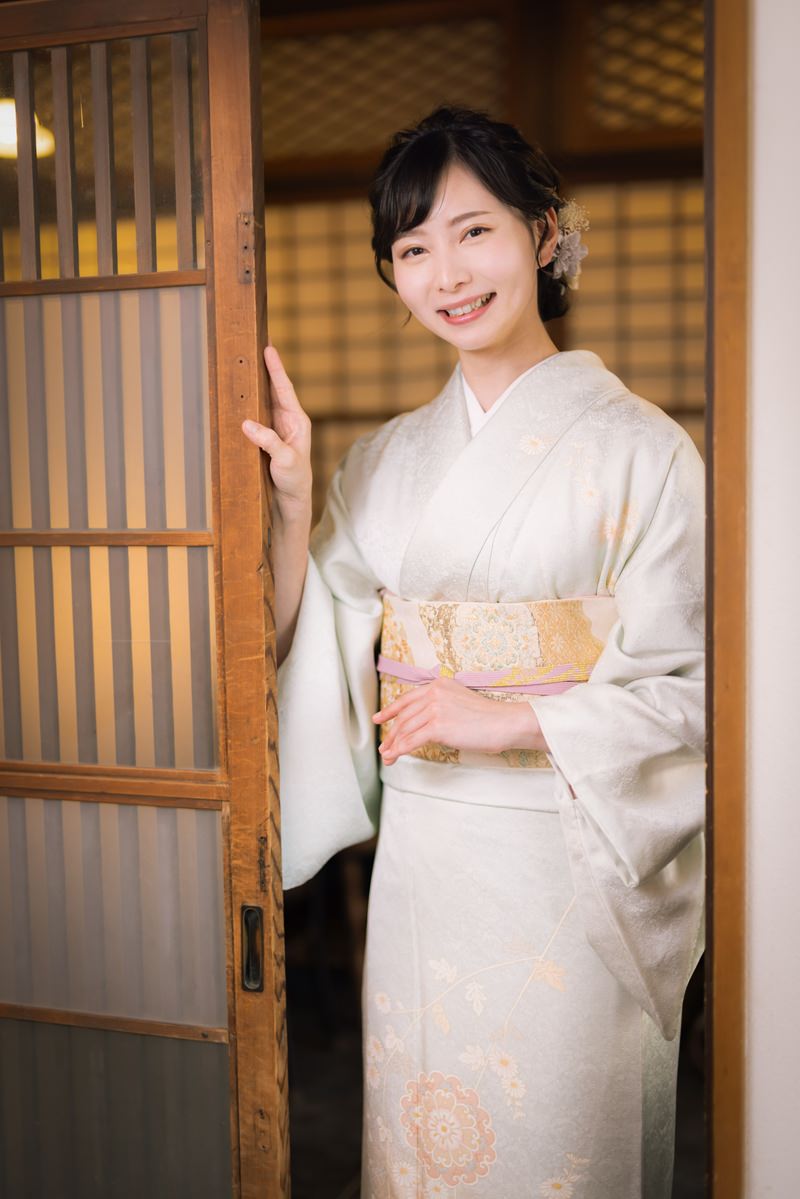 格子戸の前でクリーム色の着物を着て笑顔でお出迎えする女将［モデル：yumiko］