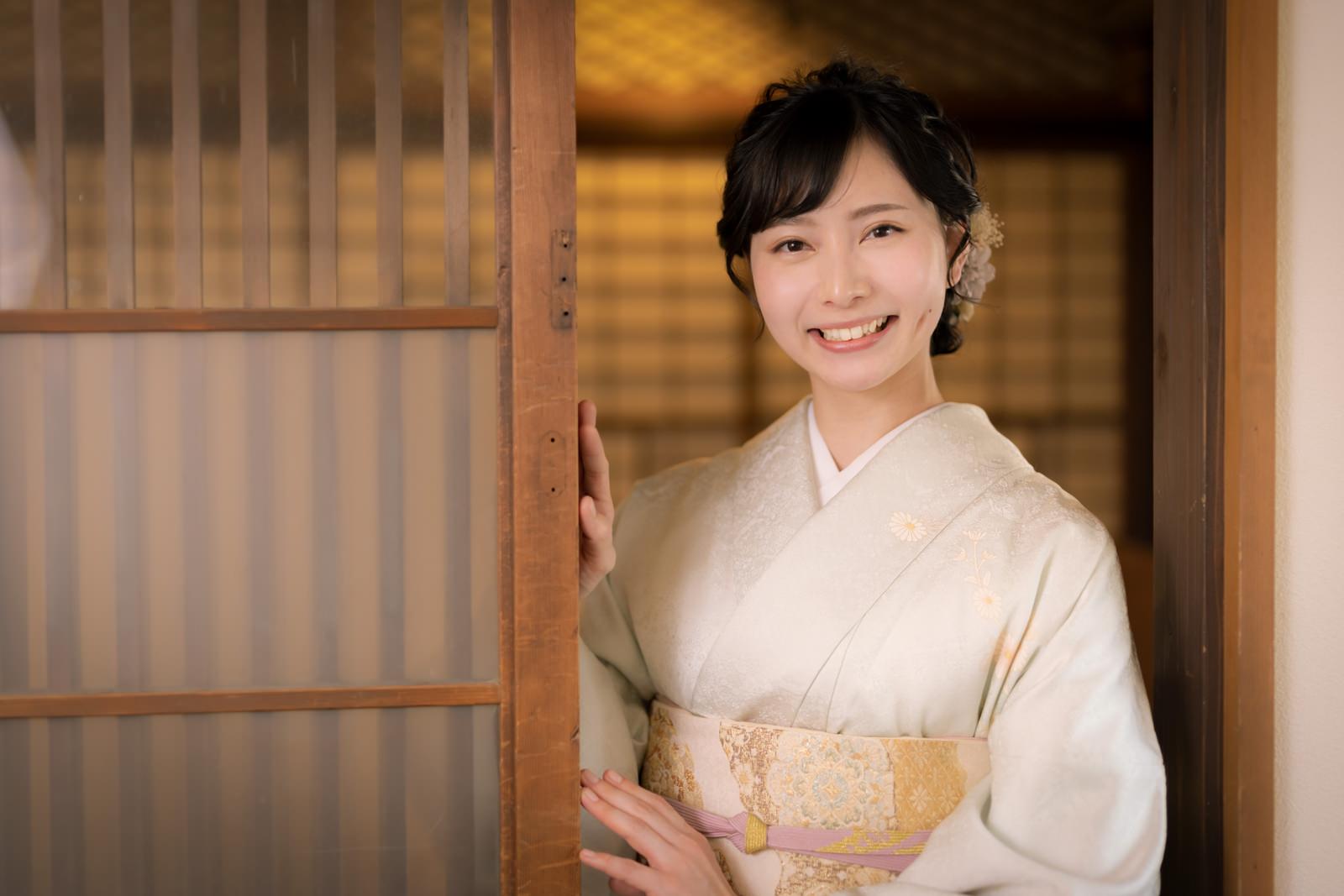 木製の格子戸の前で淡いベージュの着物を着て笑顔でお見送りをする女性［モデル：yumiko］