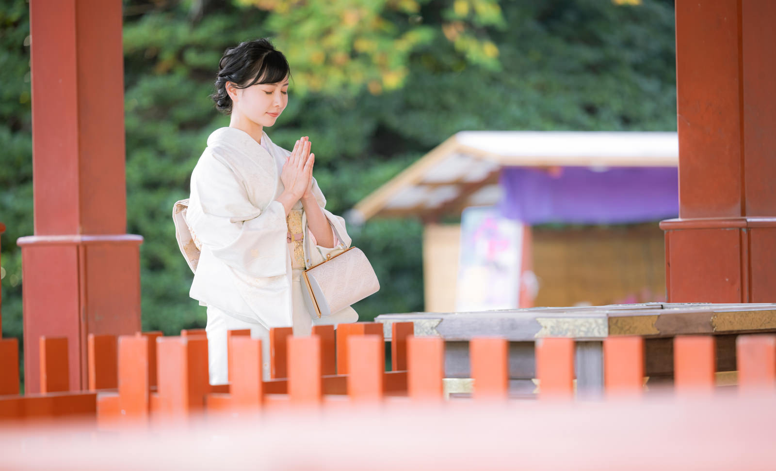神社の赤い柱の前で白い着物を着た女性が手を合わせて参拝している［モデル：yumiko］