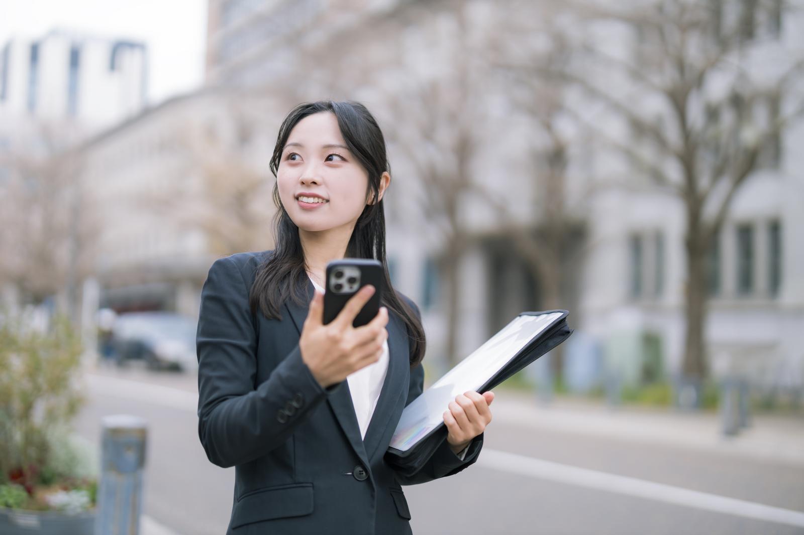 屋外でスーツ姿の若い女性2人が書類を持って笑顔で立っている［モデル：吉屋ゆり］