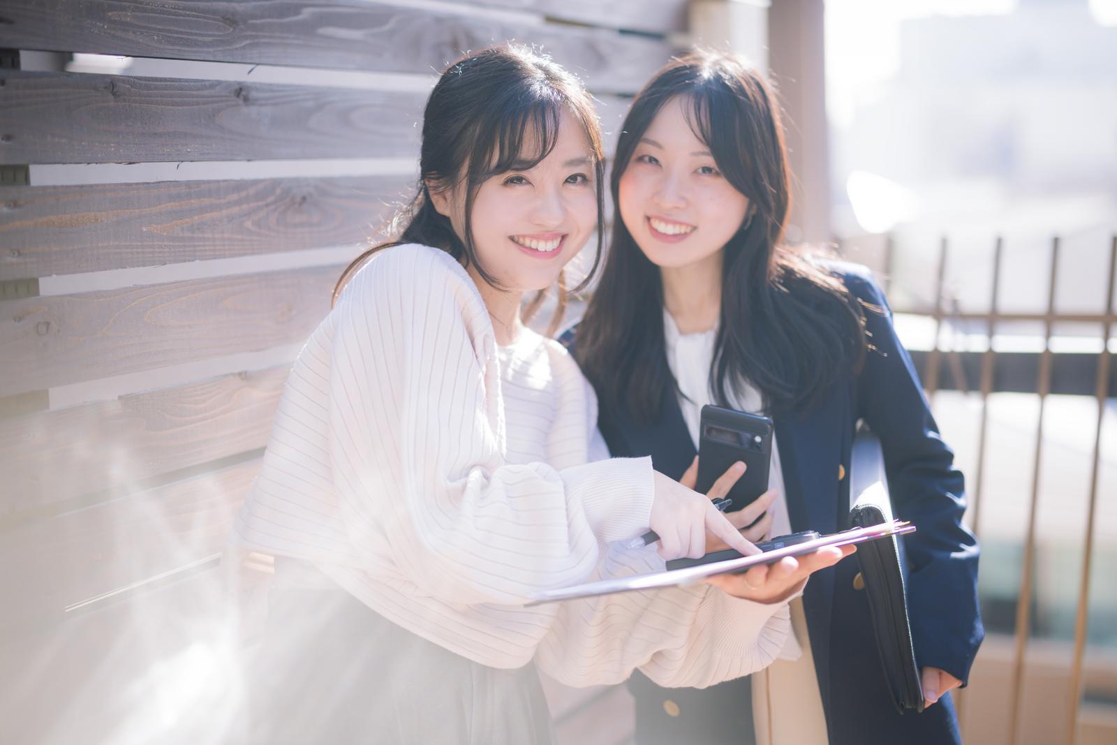 資料とスマートフォンを手に持って笑顔で提案する女性社員ふたり［モデル：吉屋ゆり 河村友歌］