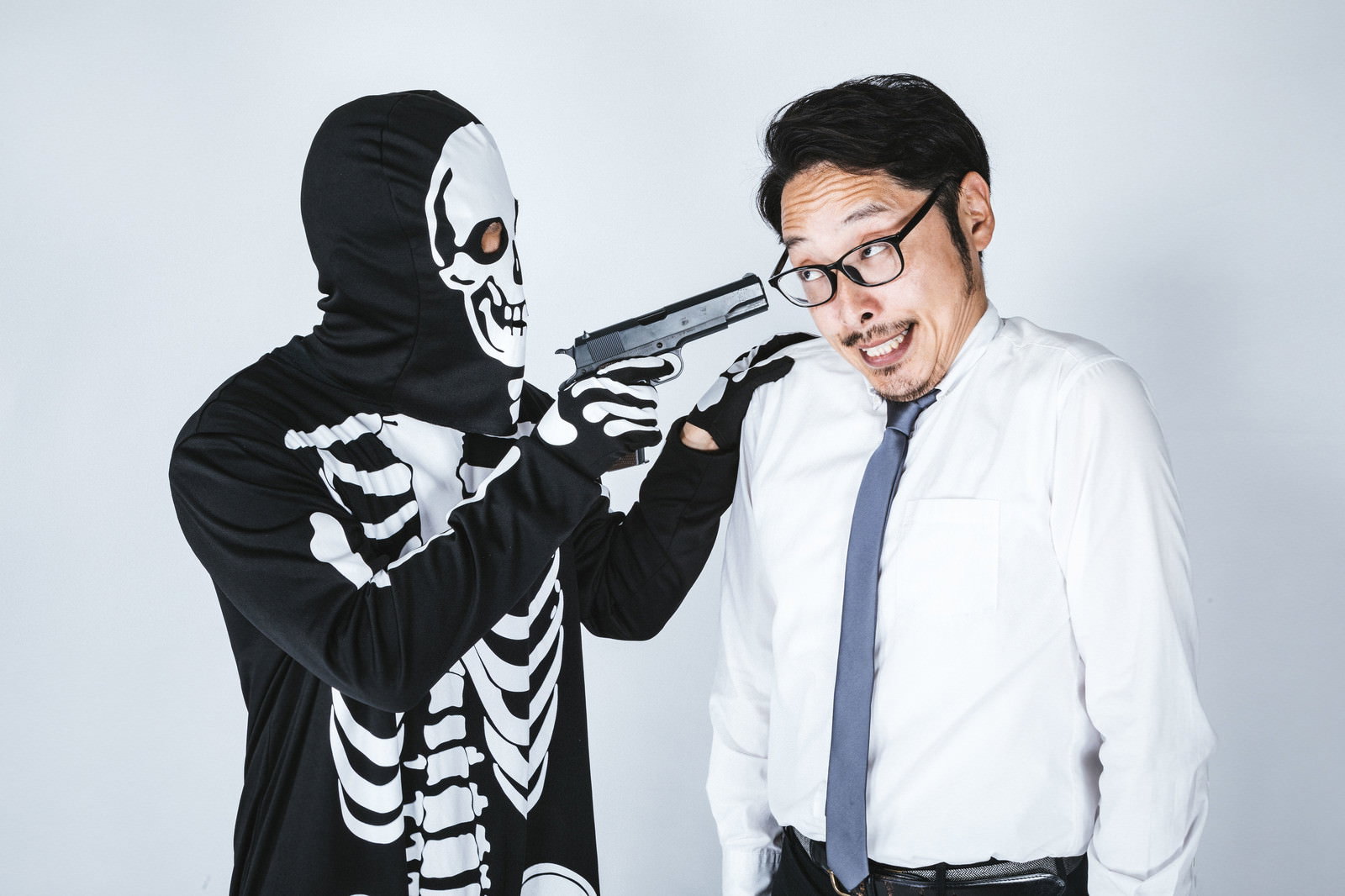 骸骨の仮装をした人物が会社員風の男性に銃を向けている様子［モデル：大川竜弥 ゆうせい］