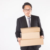 届いたダンボール箱を両手で抱えて運ぶ眼鏡の総務部長の写真
