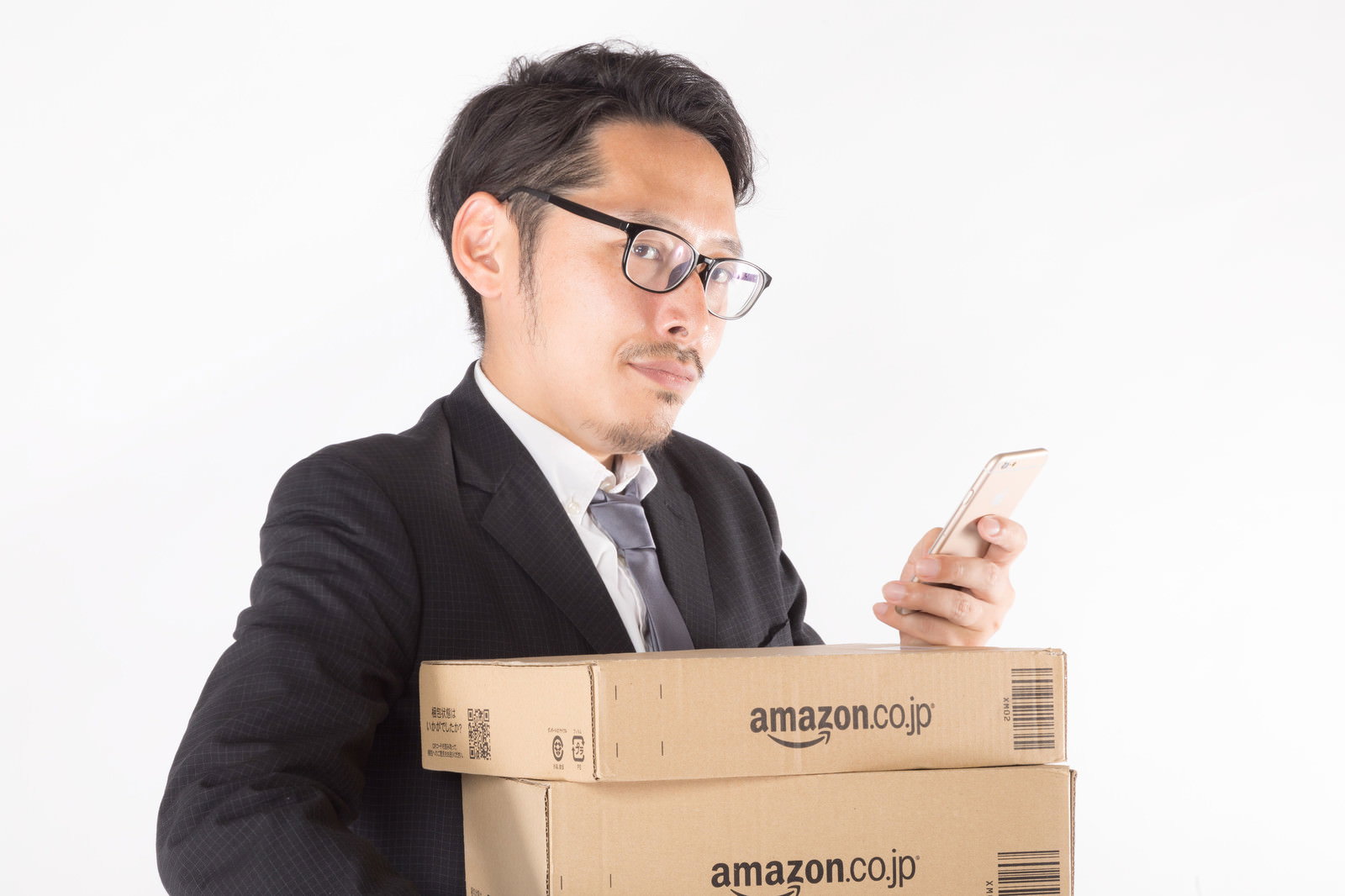 ダンボール箱の前でスマートフォンを持ち棚卸し作業をする眼鏡をかけたスーツ姿の男性［モデル：ゆうせい］