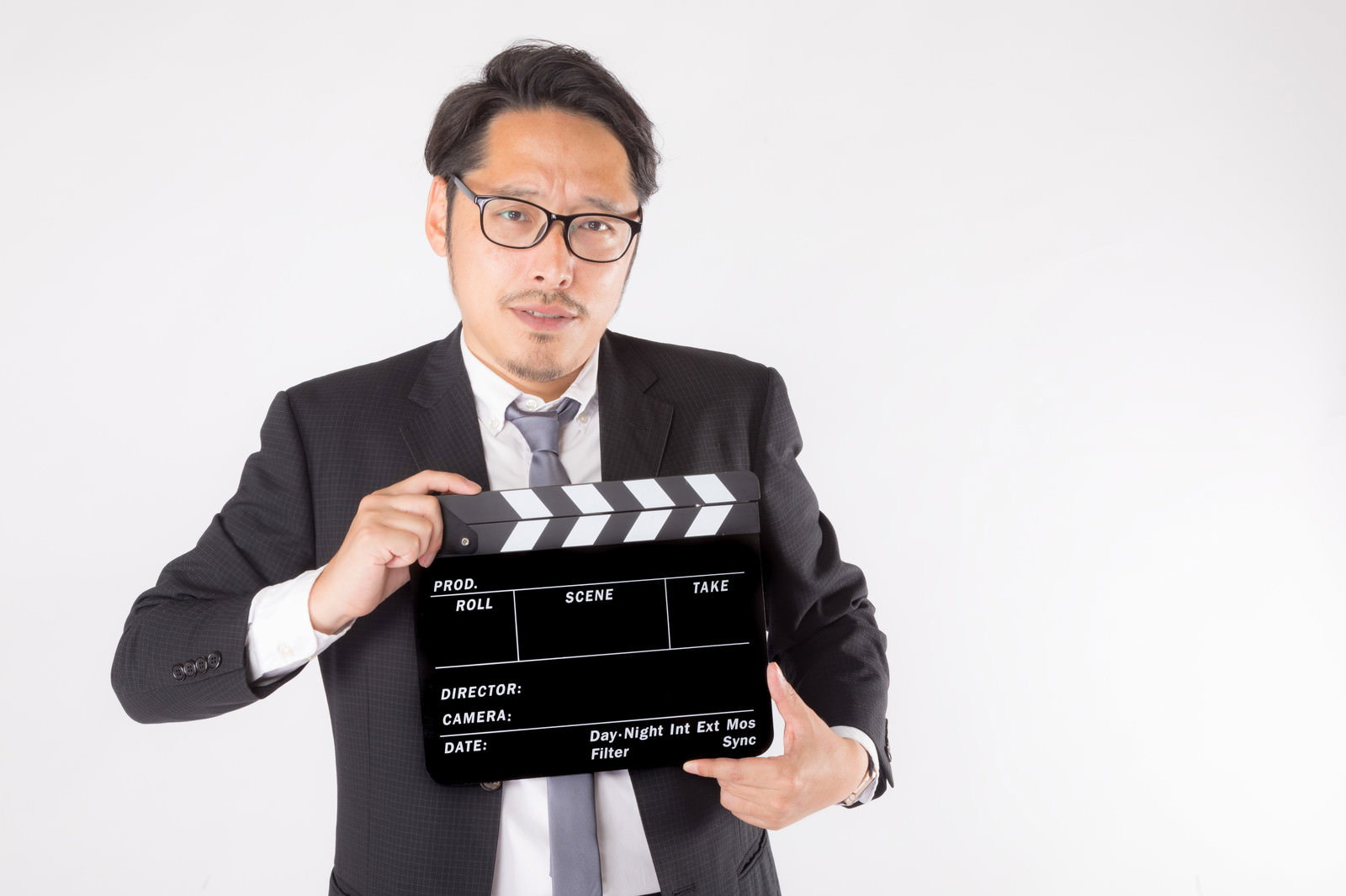 黒いスーツを着た眼鏡の男性がカチンコを持って白い背景の前に立っている［モデル：ゆうせい］
