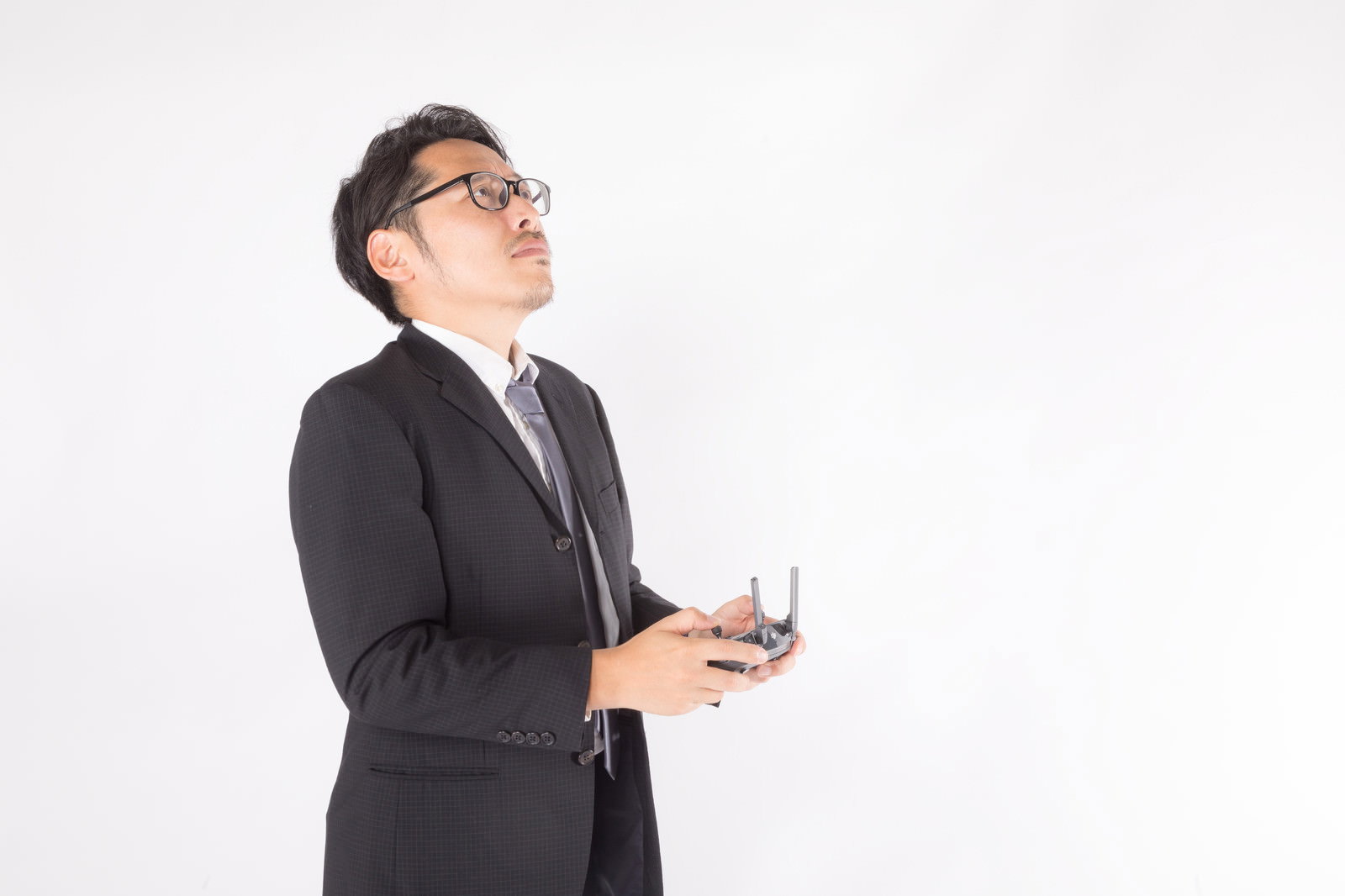 黒いスーツを着た男性が白い背景でドローンを操縦している［モデル：ゆうせい］