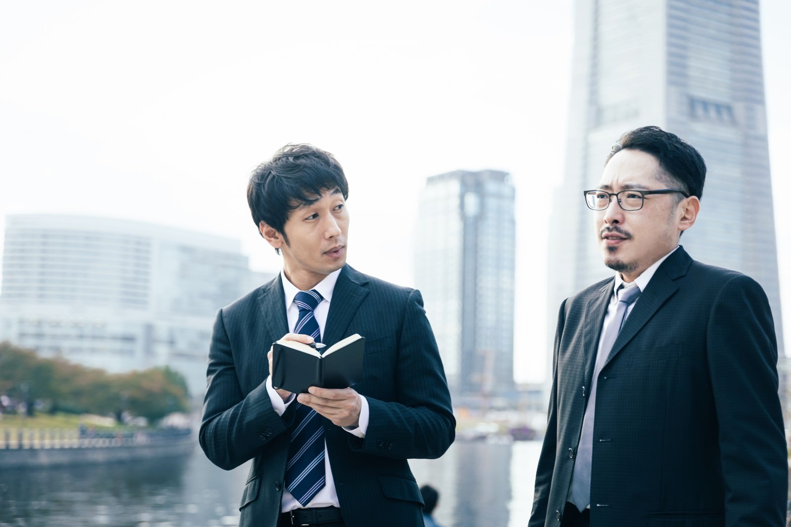 都市のビル街を背景に歩きながら取材をする新聞記者の男性2人［モデル：大川竜弥 ゆうせい］