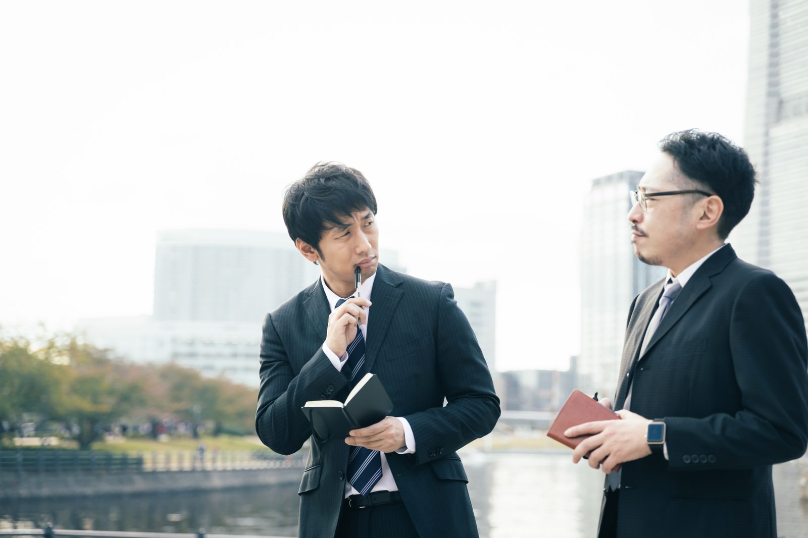 ボールペンを口元に当てて考え込むスーツ姿の男性記者［モデル：大川竜弥 ゆうせい］