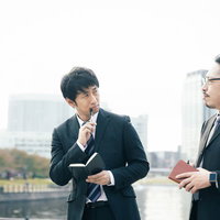 ボールペンを口に当てて考えるスーツ姿の男性記者の写真