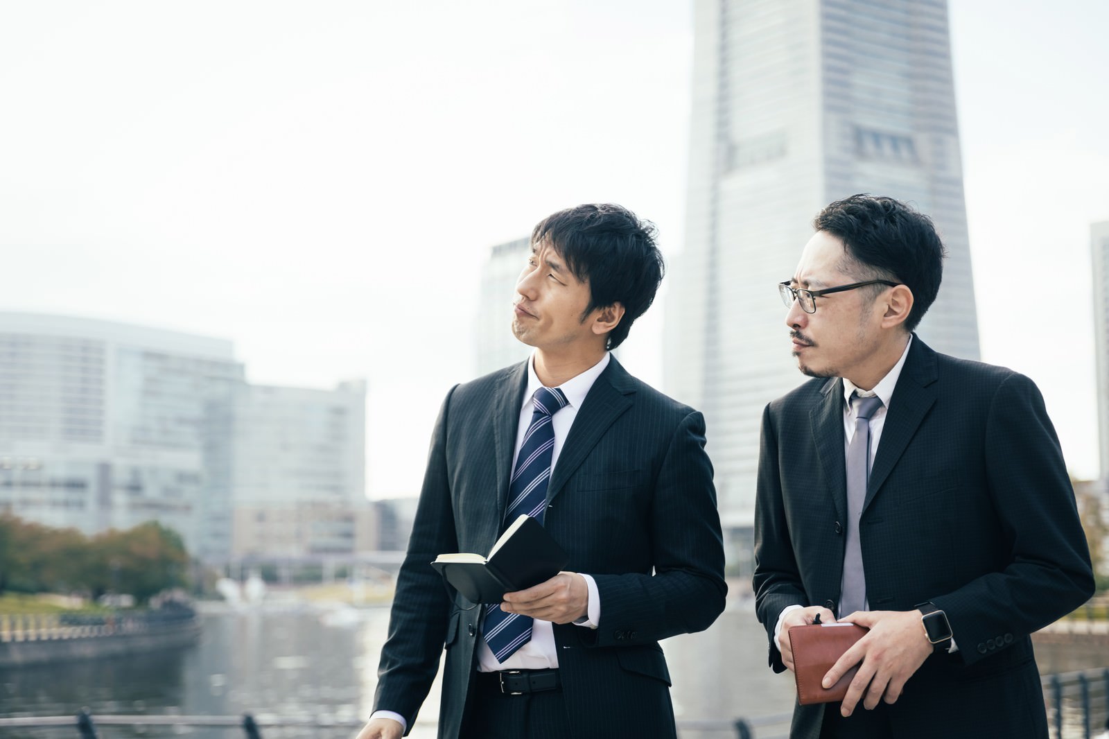 オフィスビルで書類を手に困った表情を見せるスーツ姿の二人の男性［モデル：大川竜弥 ゆうせい］