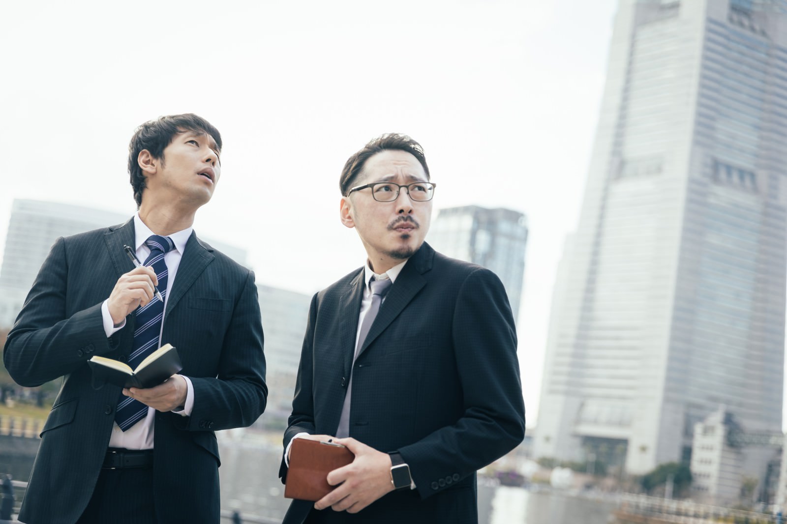 ビルの前でスーツ姿の探偵と記者が会話している写真［モデル：大川竜弥 ゆうせい］