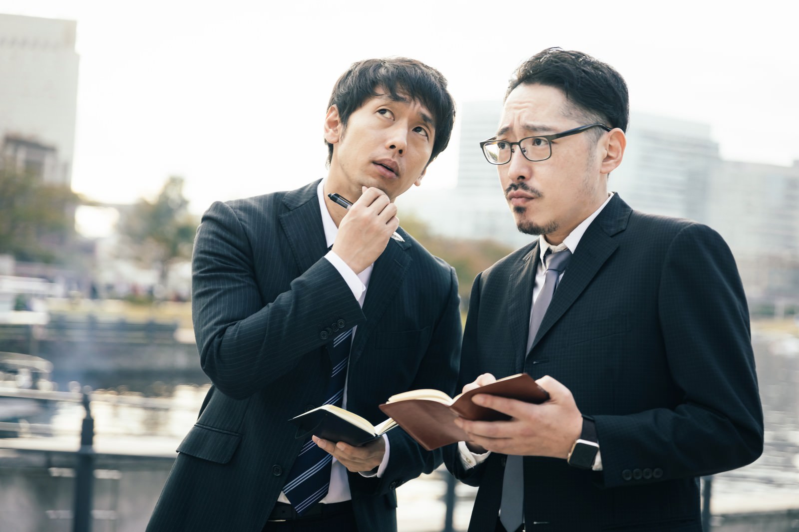 手帳を見ながら真剣に相談している2人のスーツ姿の男性［モデル：大川竜弥 ゆうせい］