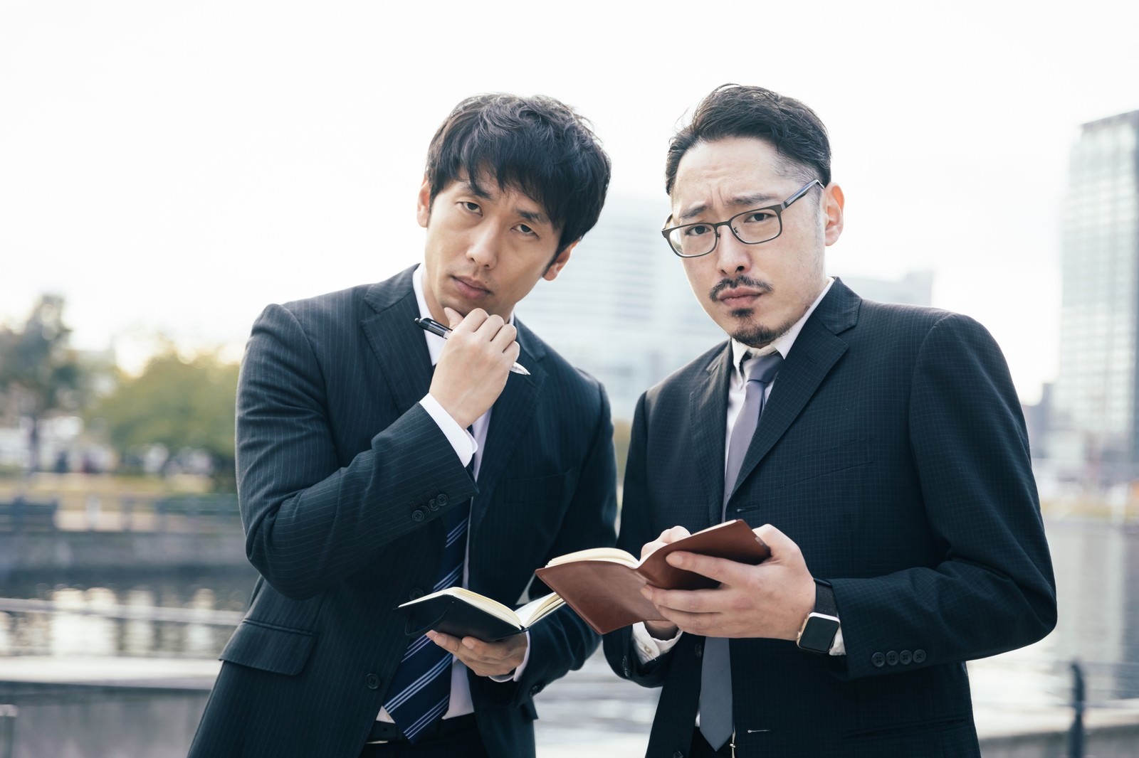 手帳を見ながら真剣に話し合う黒いスーツ姿の2人の男性［モデル：ゆうせい 大川竜弥］