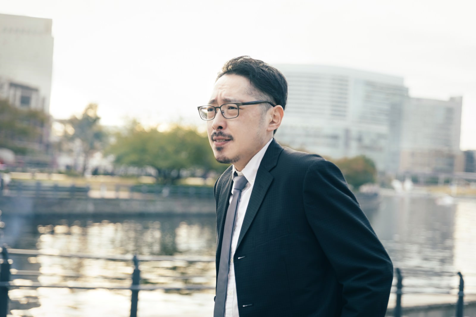 水辺の柵に立ち遠くを見つめるスーツ姿で眼鏡をかけた男性の横顔［モデル：ゆうせい］