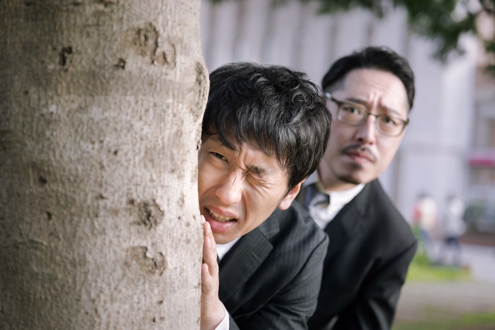 木の幹に身を隠すように立つスーツ姿の男性が驚いた表情を見せている［モデル：ゆうせい 大川竜弥］