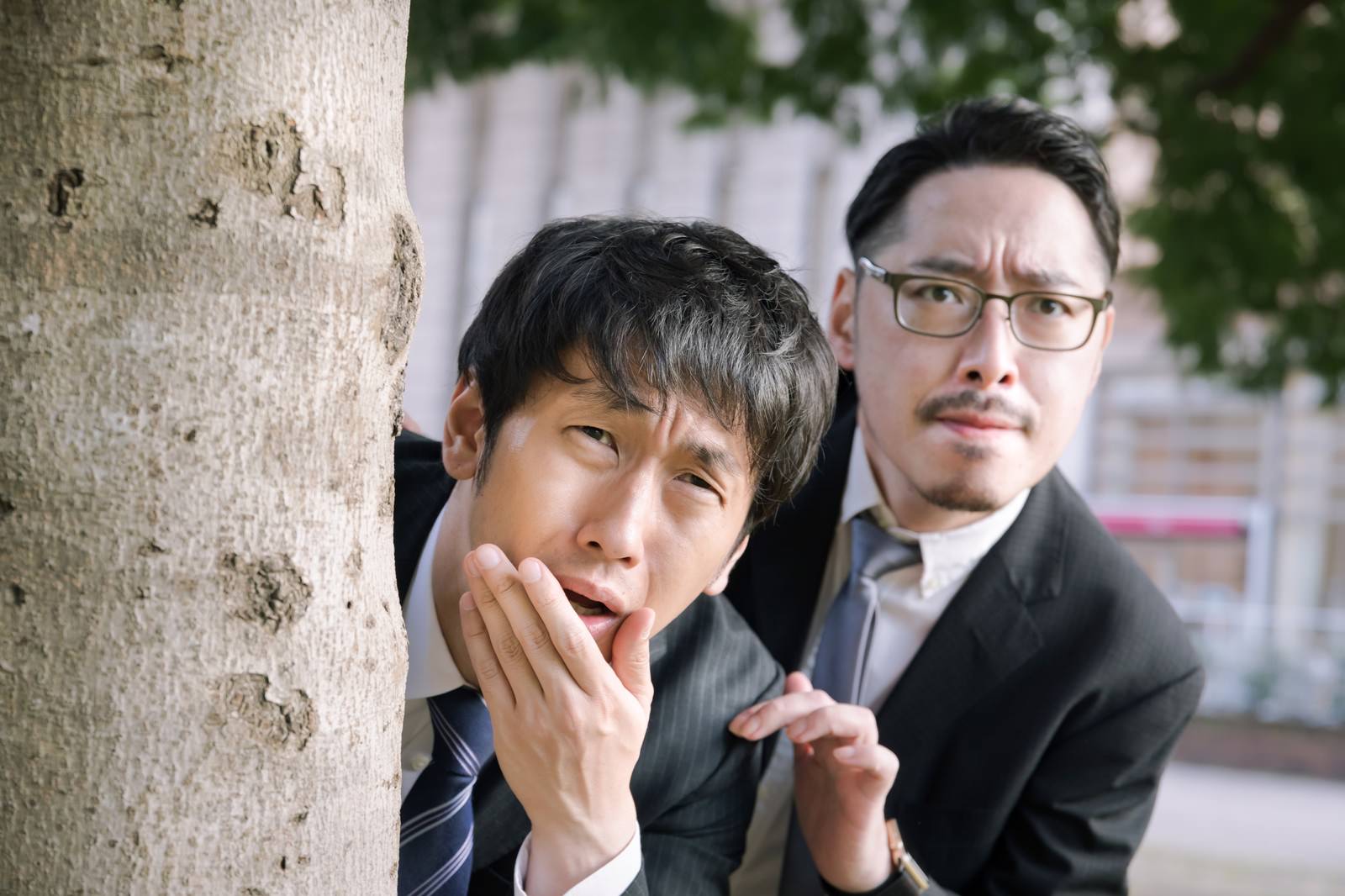 木陰に隠れながら手で口元を覆い困った表情をするスーツ姿の男性［モデル：大川竜弥 ゆうせい］