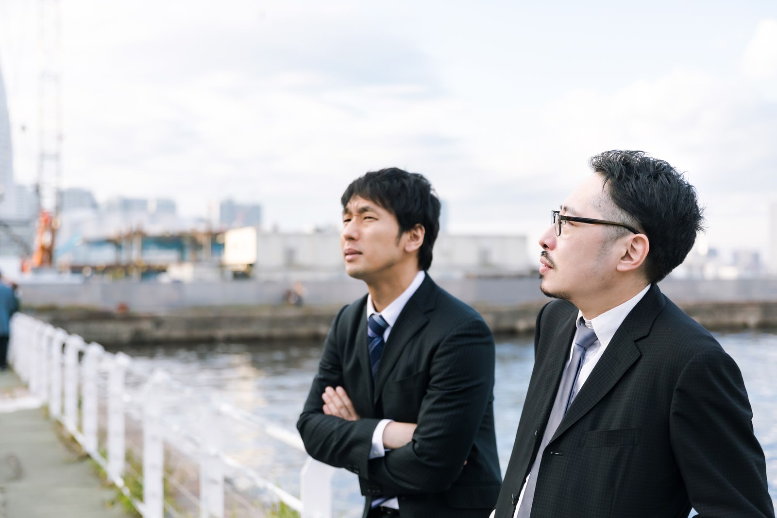 港で腕組みをして遠くを見つめるスーツ姿の男性二人［モデル：大川竜弥 ゆうせい］