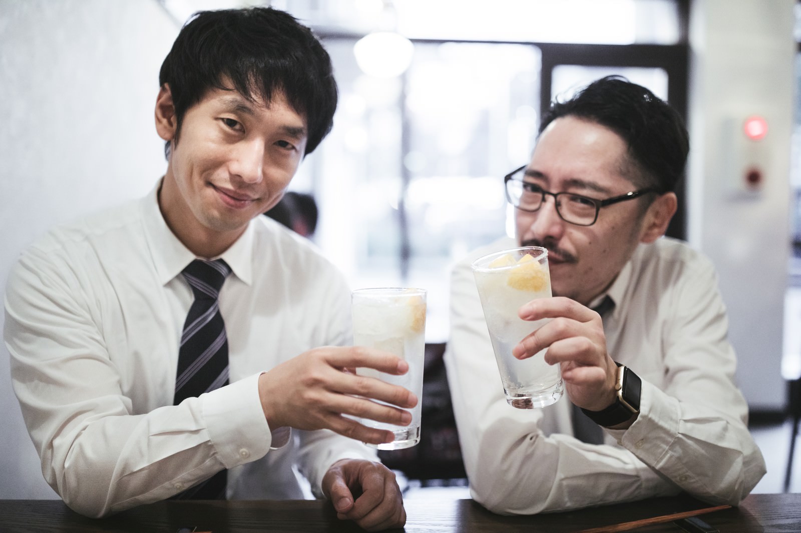 オフィスでレモンサワーを持って乾杯するスーツ姿の男性2人［モデル：ゆうせい 大川竜弥］