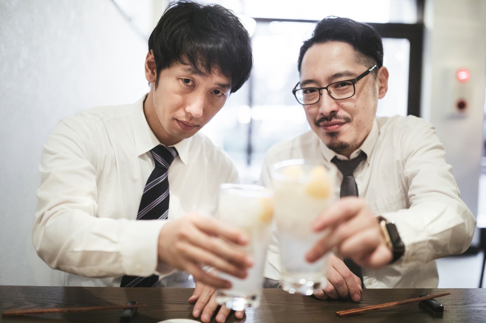 居酒屋でレモンサワーを手に乾杯するスーツ姿の男性二人［モデル：大川竜弥 ゆうせい］