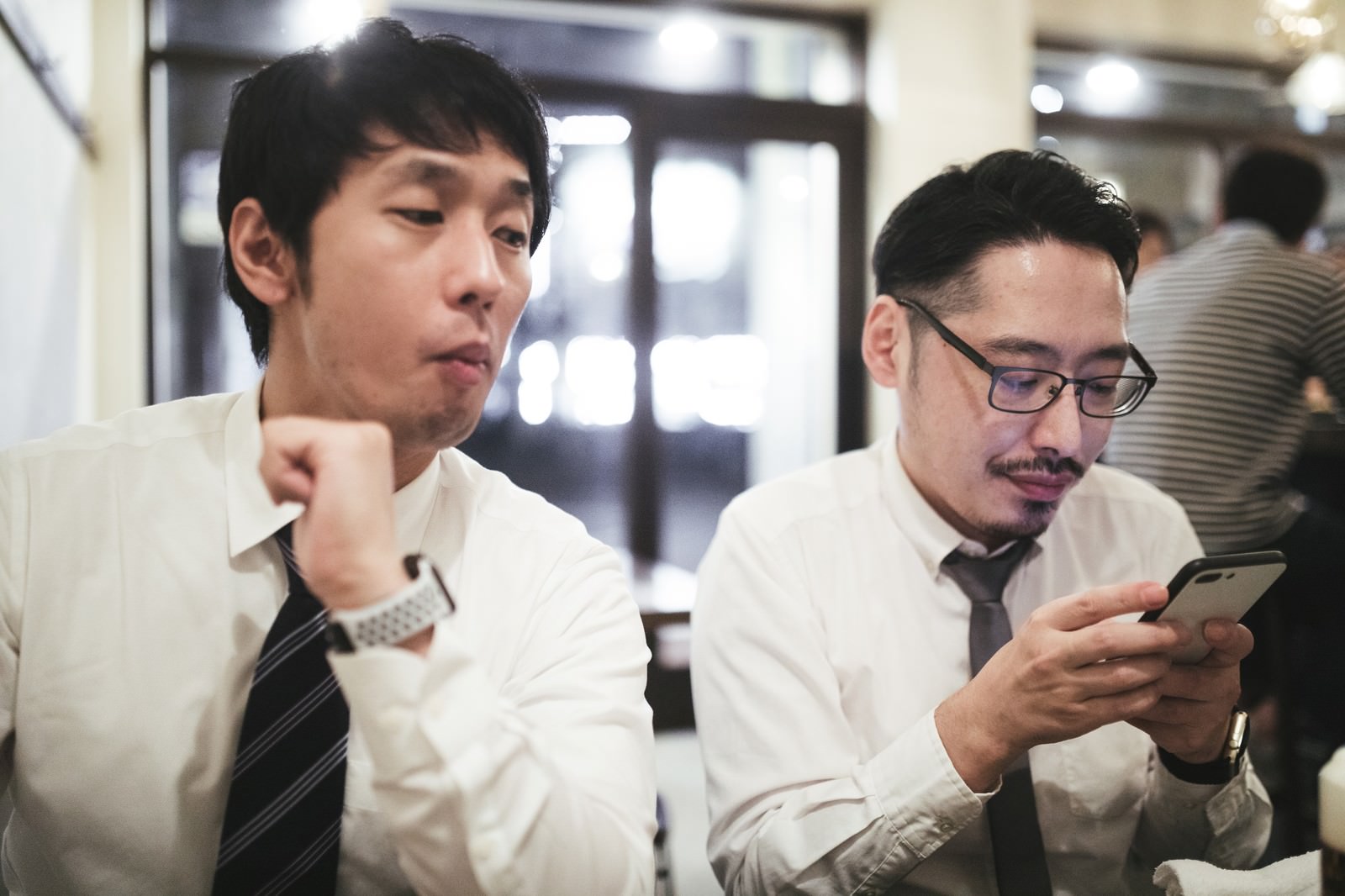 ワイシャツ姿の男性が隣の同僚のスマートフォンを横から覗き込んでいる［モデル：大川竜弥 ゆうせい］