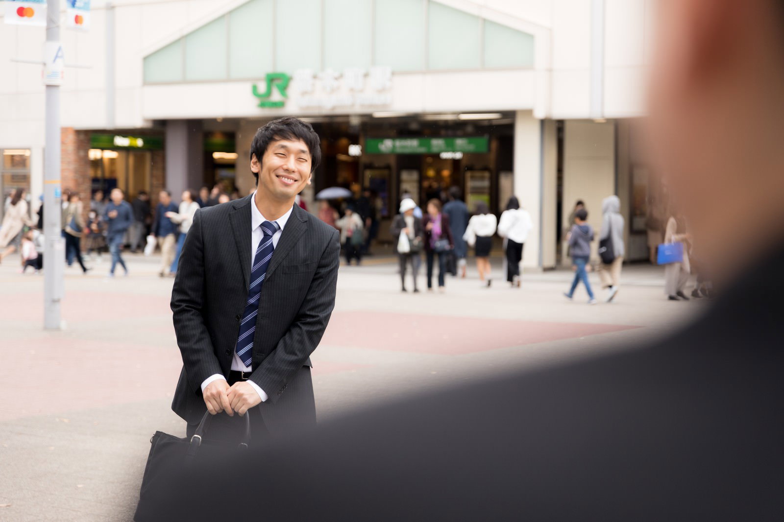 駅前の待ち合わせ場所で笑顔を見せるスーツ姿の営業マン［モデル：大川竜弥］