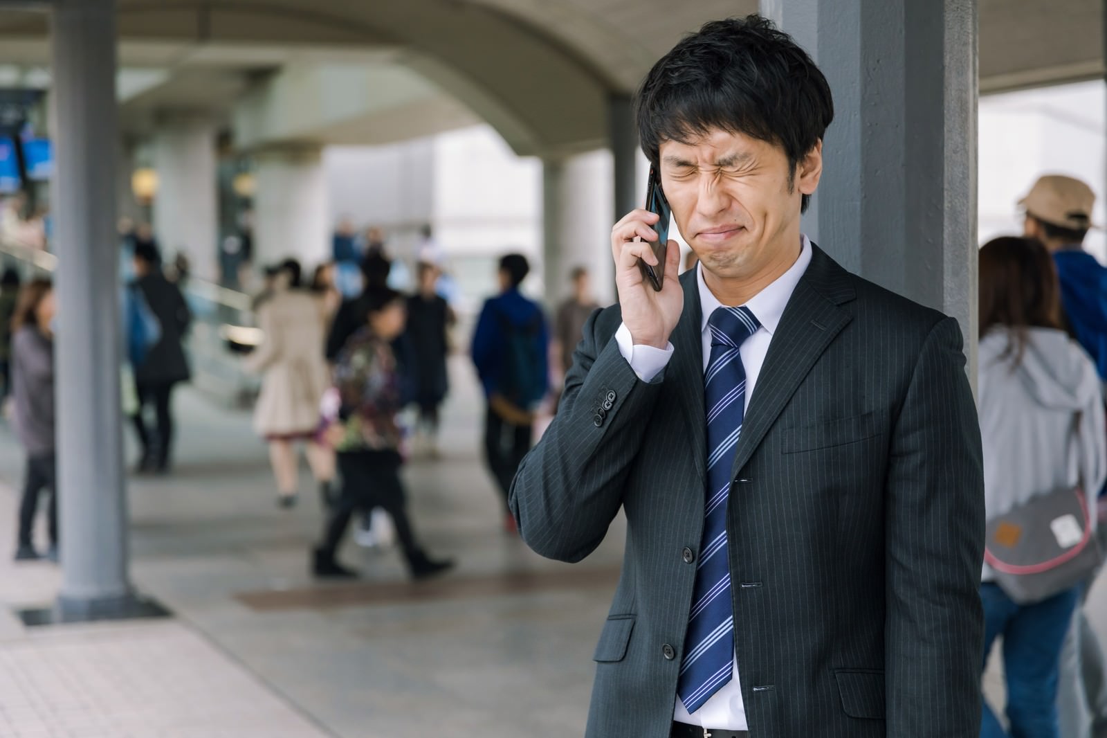 駅構内でスマートフォンで電話をかけながら悔しそうな表情を見せるスーツ姿の男性［モデル：大川竜弥］