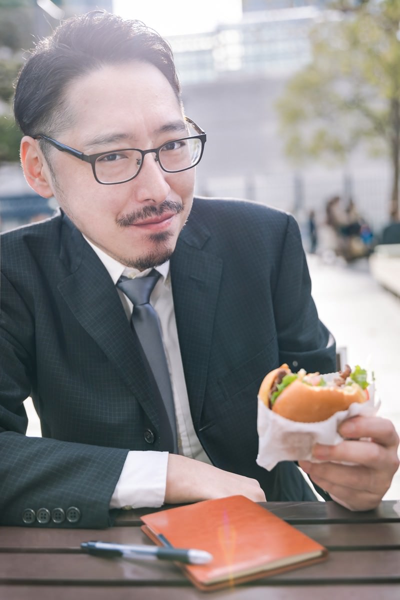 眼鏡をかけたスーツ姿の男性がテーブルでハンバーガーを食べている［モデル：ゆうせい］