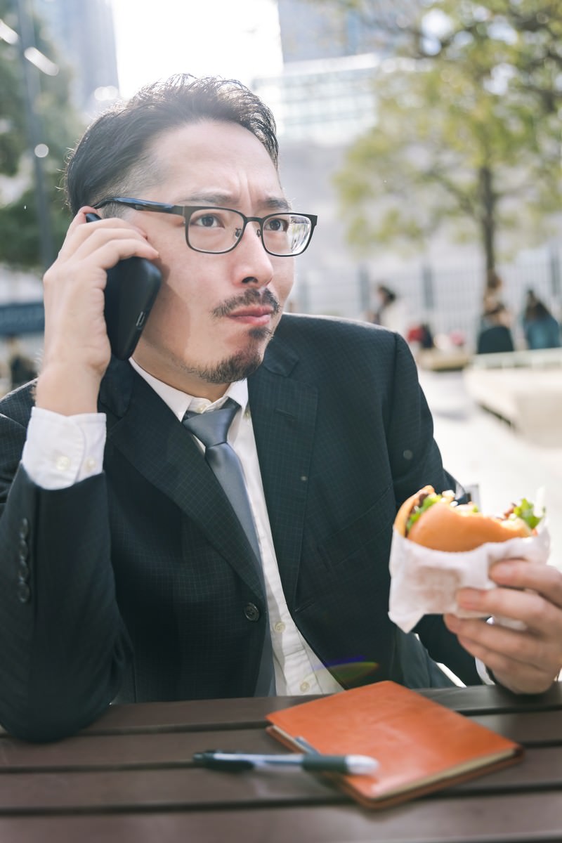 電話をしながらハンバーガーを食べるスーツ姿のビジネスマン［モデル：ゆうせい］