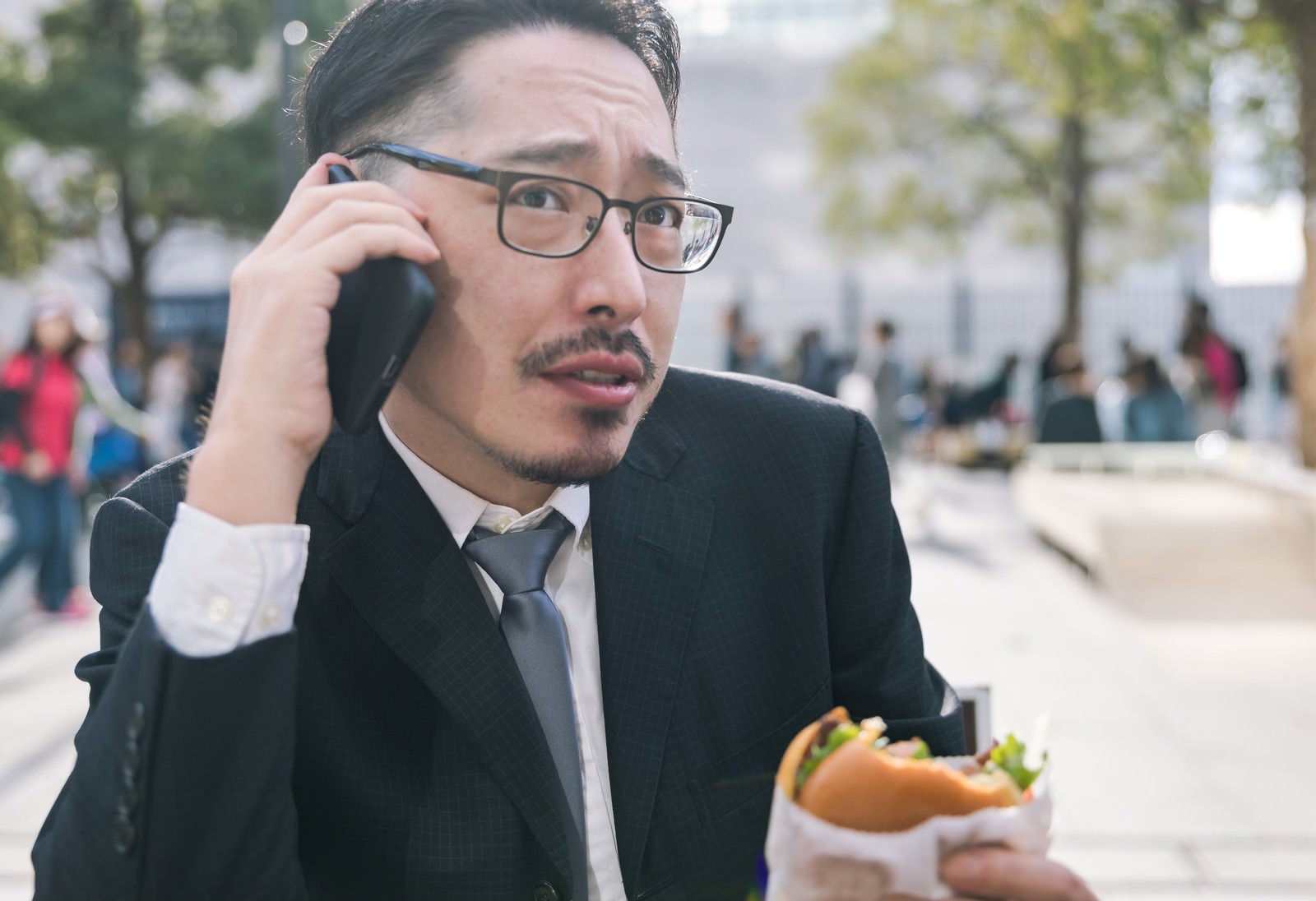 スマートフォンで通話しながらハンバーガーを持つ眼鏡をかけたスーツ姿の男性が屋外に立つ［モデル：ゆうせい］