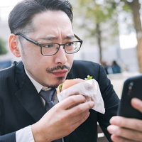 ランチタイムにハンバーガーを持ちながらスマートフォンを見る男性会社員の写真