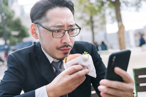 ランチタイムにハンバーガーを持ちながらスマートフォンを見る男性会社員