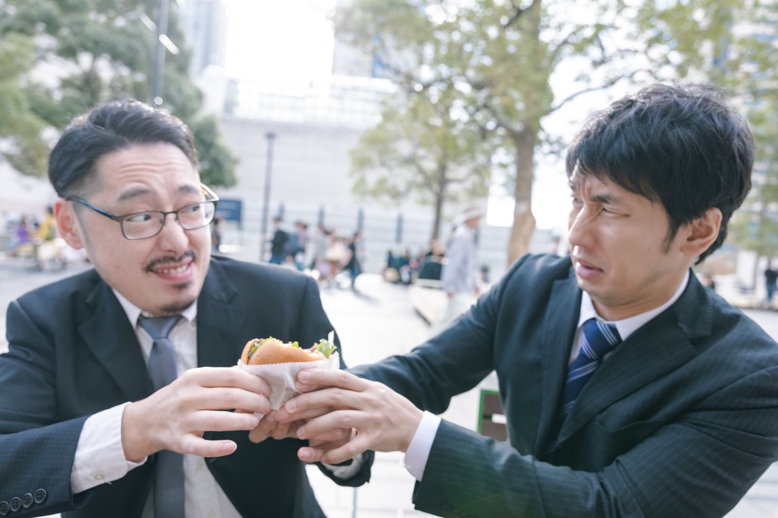 スーツ姿の眼鏡をかけた男性と若い男性がハンバーガーを取り合っている［モデル：大川竜弥 ゆうせい］