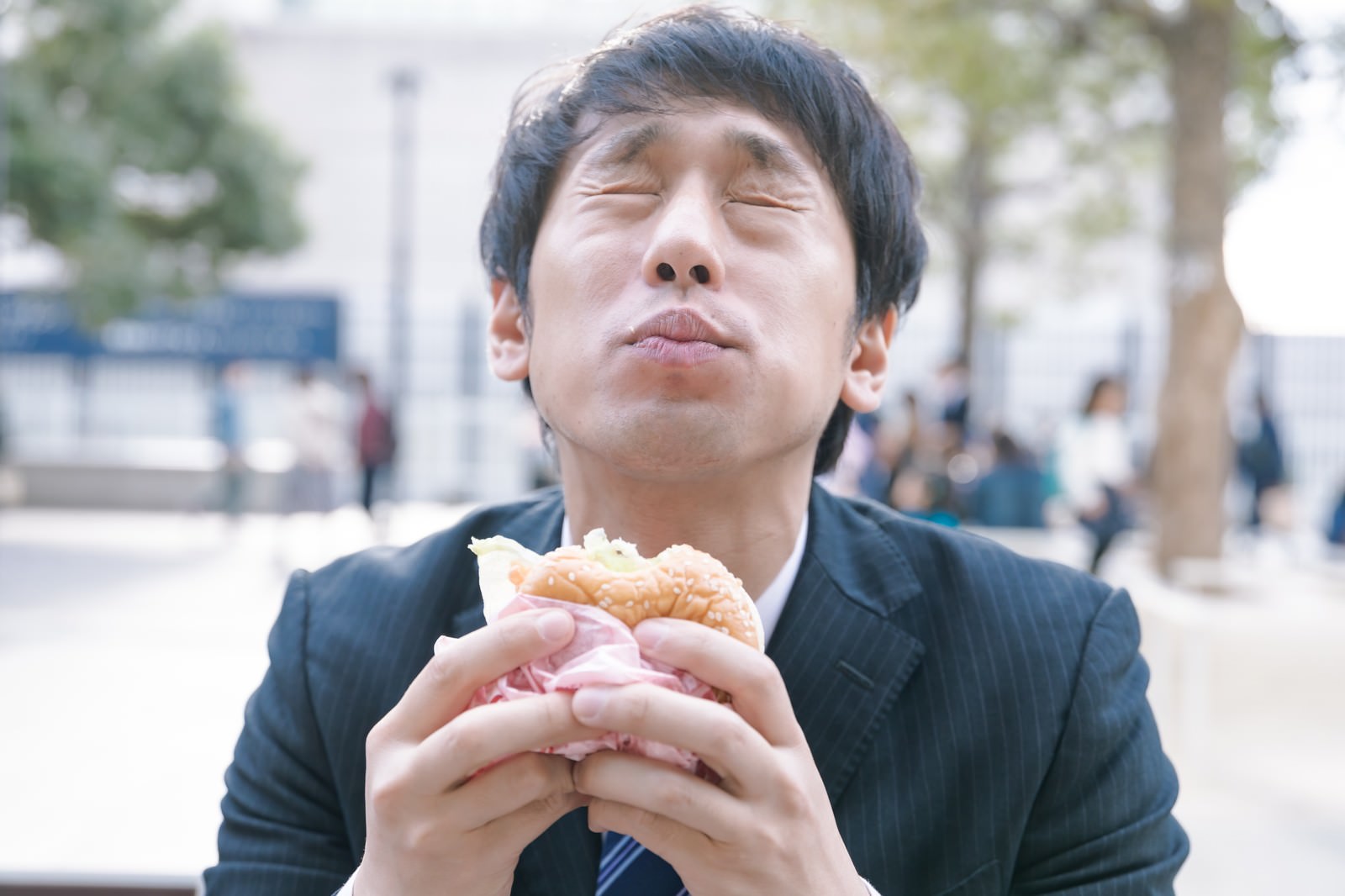 スーツを着た男性が両手でハンバーガーを持ち食べている様子［モデル：大川竜弥］