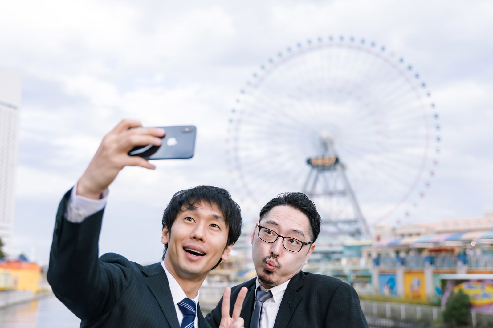 観覧車を背景にスマートフォンでセルフィーを撮る会社員男性［モデル：大川竜弥 ゆうせい］