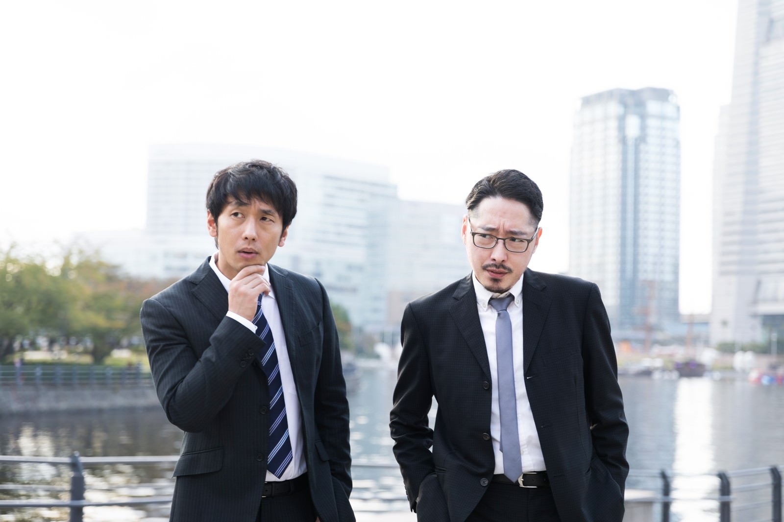 張り込み中のスーツ姿の記者とジャーナリスト2人が都市部の水辺に立つ［モデル：大川竜弥 ゆうせい］