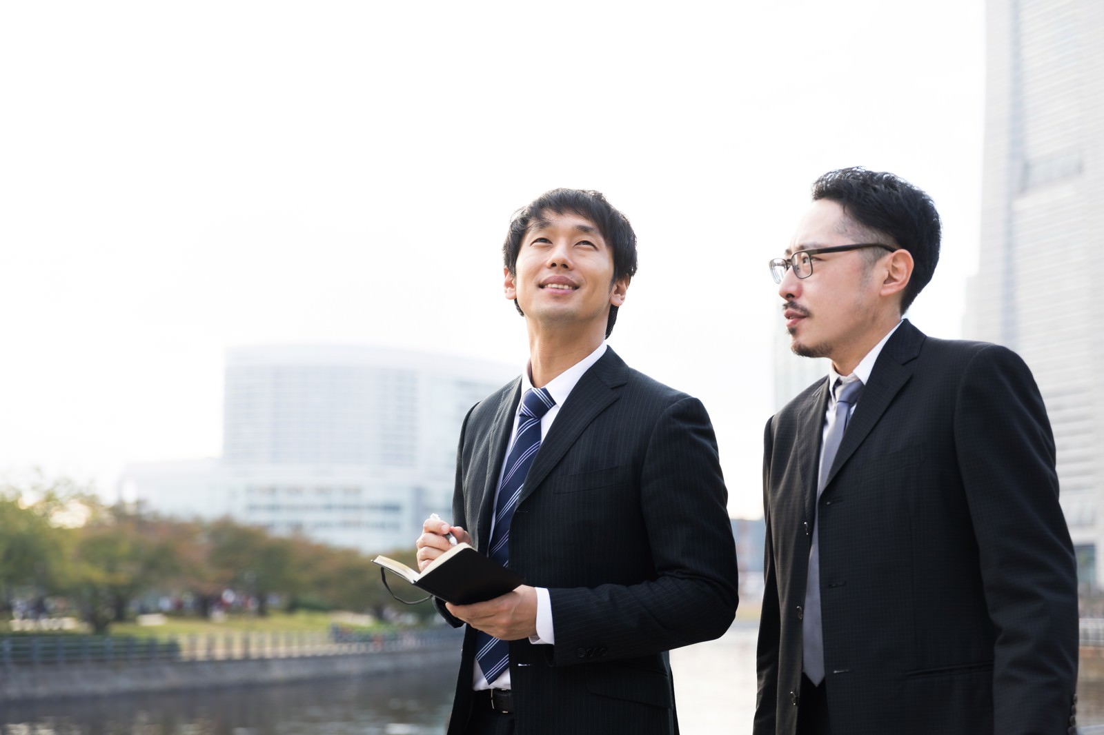 屋外で手帳を持って取材活動を行うスーツ姿の記者2人［モデル：大川竜弥 ゆうせい］