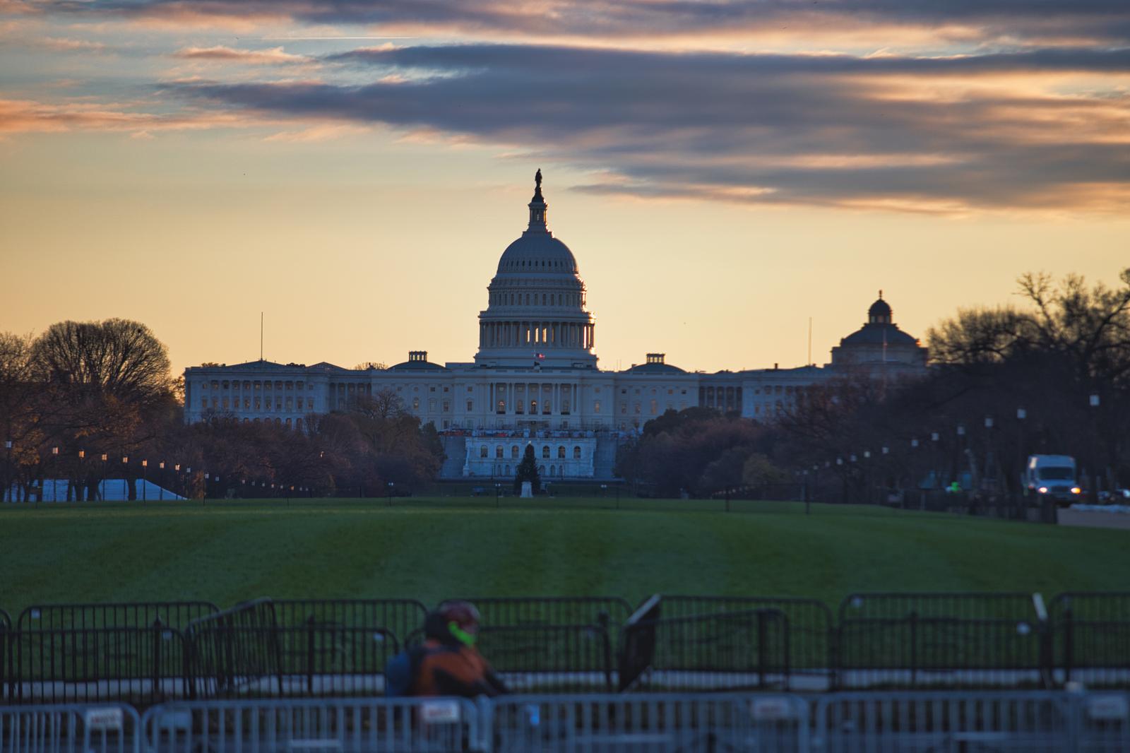朝焼けに照らされるアメリカ国会議事堂のドーム。奥に白い建物が見え、上空には橙色と青紫色の雲が広がっている。