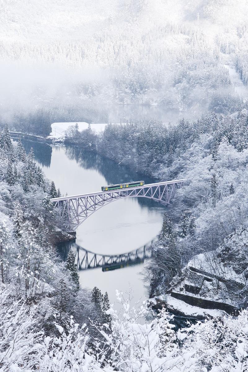 The winter landscape of a forest and river covered in white snow, with a yellow-green train running across a steel bridge.